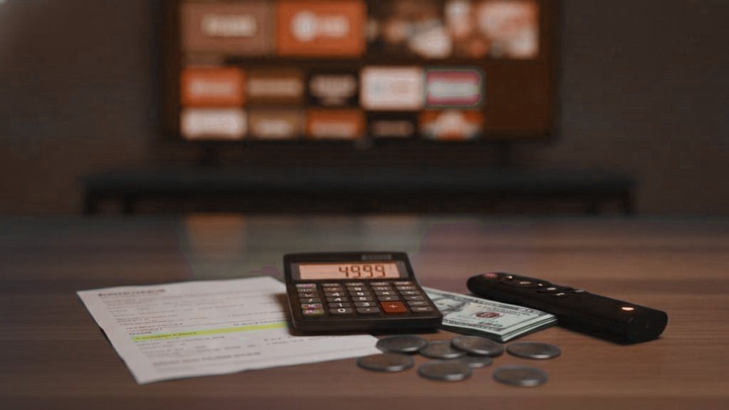 A calculator, dollar bills, coins, a streaming stick and a remote on a table in front of a TV