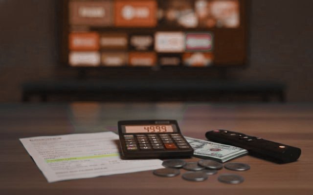 A calculator, dollar bills, coins, a streaming stick and a remote on a table in front of a TV
